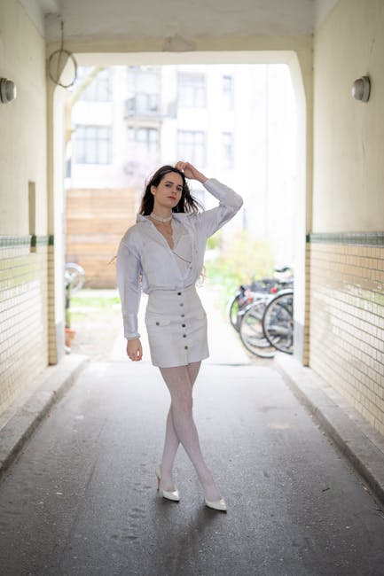 A young woman with long, dark hair stands in a dimly lit underground passageway, with pale yellow walls and a tiled bottom. She is dressed in a white blouse with a V-neck and long sleeves, tucked into a high-waisted, beige button-front skirt. The skirt features three rows of dark buttons running vertically down the front, and she wears sheer white tights and white high heels. Her right hand is raised to touch her head, and she gazes to the side with a neutral expression. In the background, outside the tunnel, bicycles are visible leaning against a wooden fence, and a glimpse of a residential building with multiple windows can be seen, indicating an urban environment. The natural light from the outside contrasts with the shadowed interior, highlighting her figure and outfit, creating a balanced composition that emphasizes her presence in the space. Although the setting appears casual, her polished appearance and the neutral surroundings subtly evoke themes of private or on-site clearance, aligning with the context of independent rubbish removal or waste handling. The scene is focused on her attire and the contrasting environment, with no visible rubbish or waste objects present.