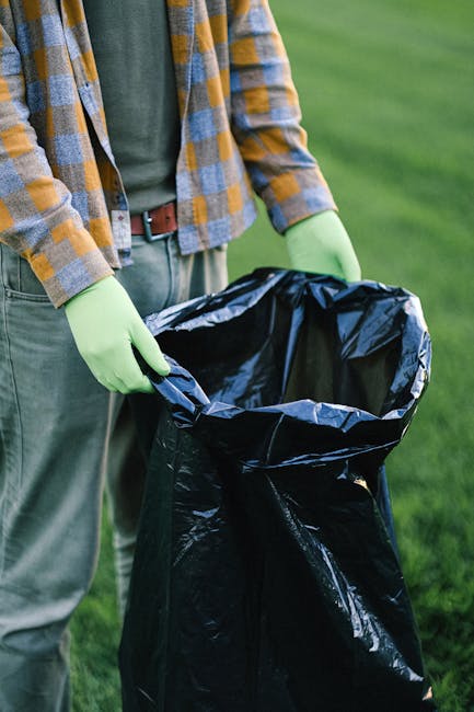 A person standing outdoors on a grassy area is holding open a large black plastic rubbish bag with both hands, which are protected by bright green gloves. The individual is dressed in casual clothing, wearing a multicolored plaid shirt over a dark T-shirt, with a brown belt and grey trousers. The background consists of a well-maintained green lawn, with no other objects or structures visible. The scene suggests an activity related to waste collection, possibly an on-site rubbish removal or garden clearance, reflecting private waste management or alternative rubbish disposal methods typical of independent rubbish removal services like those offered in Colliers Wood. The focus on the open bag and the gloves emphasizes an organised approach to rubbish handling, aligning with environmentally responsible waste collection practices.