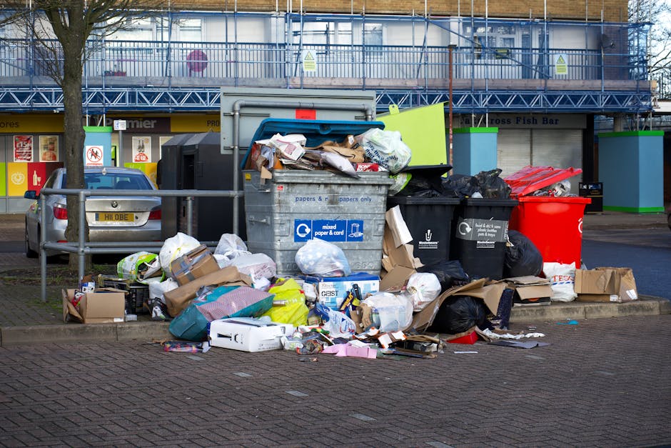 A collection of overflowing mixed waste and rubbish bags, including cardboard boxes, plastic bags, and loose paper, piled around a large grey metal recycling bin on a paved urban street. The waste appears to be a combination of household and possibly commercial refuse, with some items partially spilling onto the pavement. The grey bin is marked with blue and white labels indicating it is for mixed paper and card, and its lid is open, revealing crumpled paper and packaging materials inside. To the left of the bin, a small silver car is parked beside a barrier railing, with a tree and commercial storefronts in the background, one with a yellow sign and another with a blue facade under a building with scaffolding and safety netting. The scene suggests an area where private rubbish collection services might handle waste removal, contrasting with standard municipal disposal, and emphasizes the importance of professional rubbish removal services such as those offered by [COMPANY_NAME]. The overall environmental setting and waste arrangement highlight typical urban rubbish accumulation during periods of insufficient collection or spillover from collection services.
