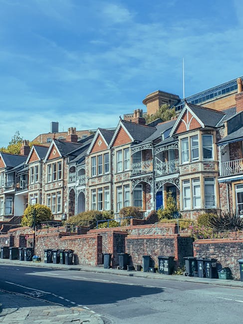 A row of ornate Victorian-style terraced houses with decorative ironwork on the balconies and bay windows, situated along a slightly inclined street with a brick retaining wall and a series of black wheelie bins lined up at the base. The houses are constructed with brick and painted finishes, featuring gabled roofs, small front gardens with shrubs, and a clear blue sky overhead. The scene reflects a residential area with individual property features, typical of local urban environments where independent waste collection and on-site rubbish removal might be required, managed by a professional waste management company like rubbishcollectioncollierswood.co.uk.